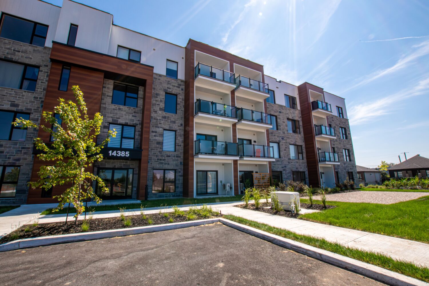 Vue d'ensemble de la façade d'un immeuble d'appartements urbain à louer. Le bâtiment moderne présente une combinaison de matériaux, incluant du revêtement blanc, de la maçonnerie de pierre grise et des accents de bois. On peut voir plusieurs balcons avec des garde-corps en verre et métal noir. Un stationnement et des aménagements paysagers avec de la pelouse et des arbustes sont au premier plan. L'adresse "14385" est visible près de l'entrée.