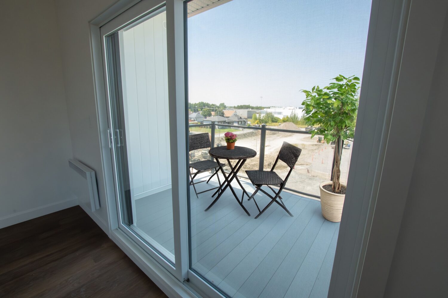 Vue d'un balcon meublé, accessible par une grande porte patio en verre. Le balcon, avec son plancher gris, est aménagé avec une petite table ronde noire et deux chaises assorties, ainsi qu'une plante en pot et une petite jardinière de fleurs sur la table. Le garde-corps est en verre et en métal noir, offrant une vue sur un quartier résidentiel et un chantier de construction.