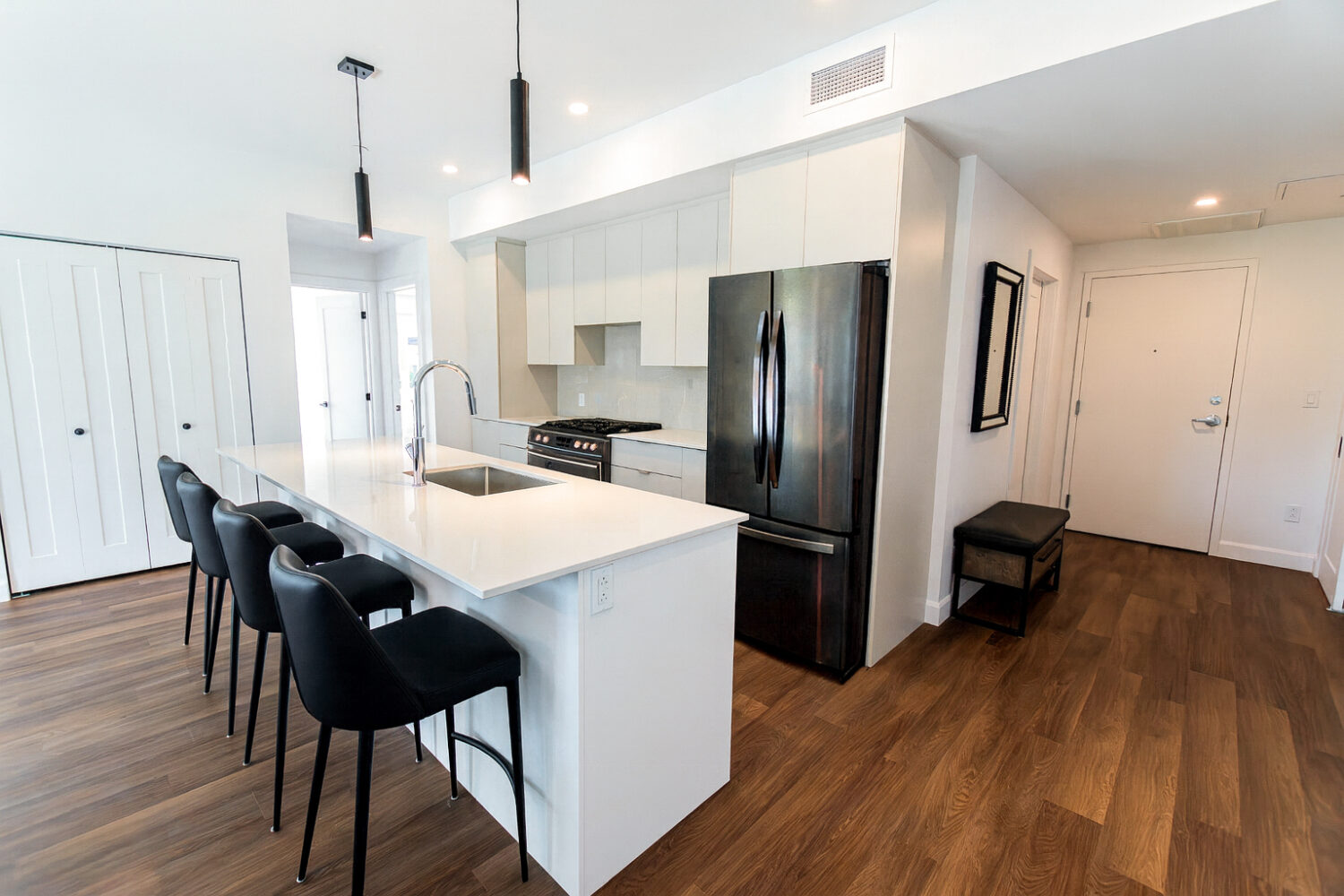 Vue d'une cuisine moderne dans un appartement. On y voit un grand îlot de cuisine blanc avec un évier, un robinet et quatre tabourets de bar noirs. La cuisine est équipée d'électroménagers noirs, d'armoires blanches et d'un dosseret de tuiles blanches. Un plancher de bois s'étend de la cuisine jusqu'à l'entrée, où se trouvent un petit banc et un miroir encadré de noir.