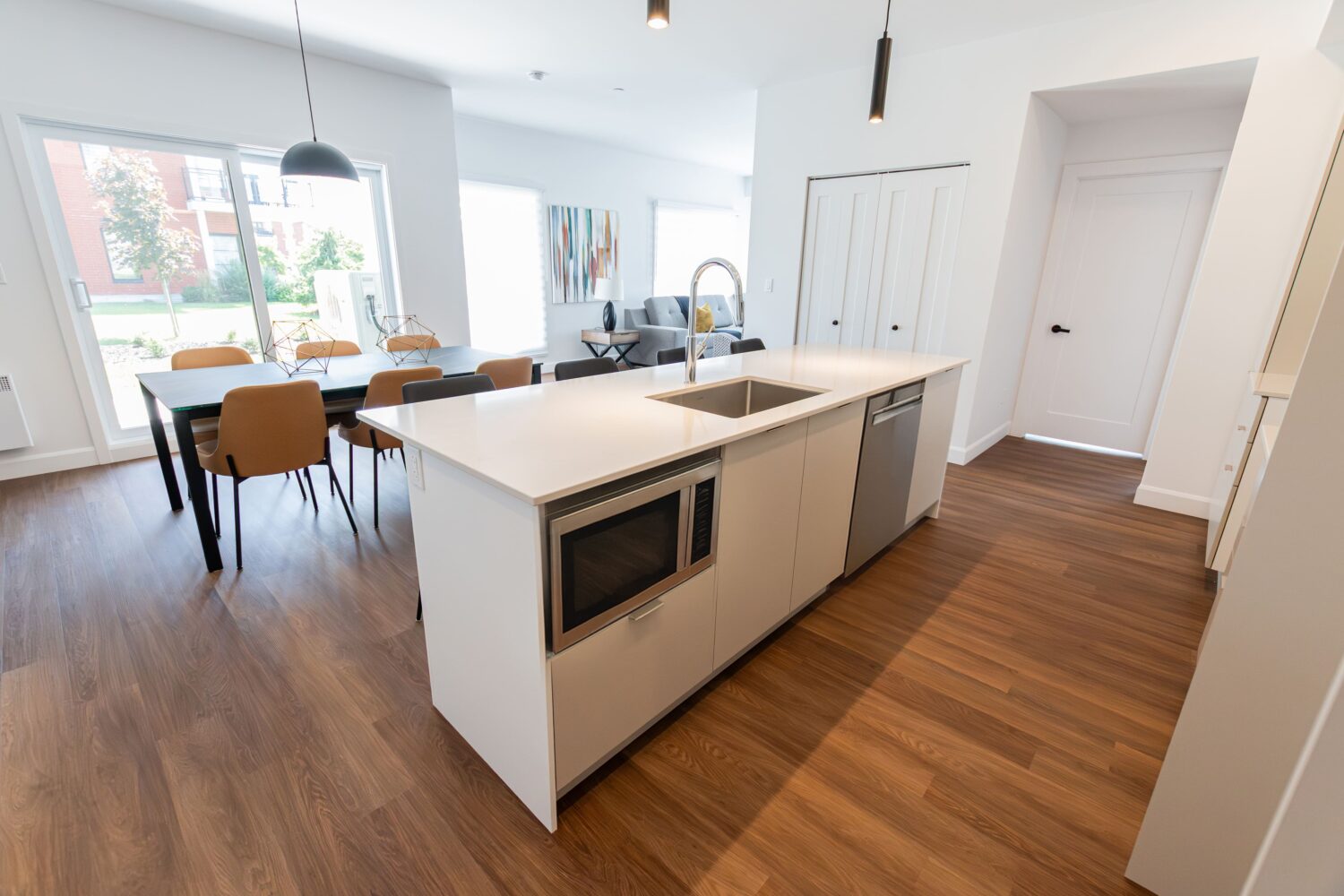 Vue d'une cuisine à aire ouverte dans un appartement moderne. Un grand îlot blanc avec un évier, un robinet et un four à micro-ondes intégré est au premier plan. Derrière l'îlot, on aperçoit une salle à manger avec une table noire et des chaises noires et brunes, menant à une porte patio et un balcon. Le plancher en bois s'étend de la cuisine à la salle à manger, avec un salon visible au loin.