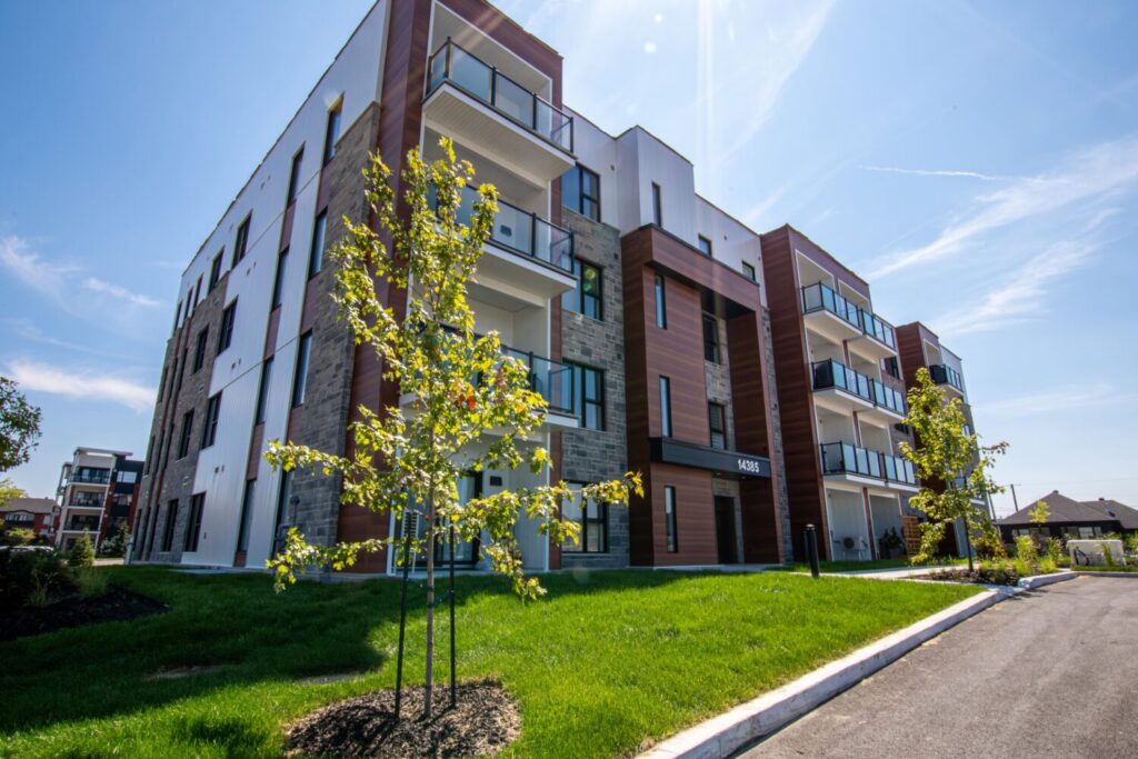 Vue de l'extérieur d'un immeuble d'appartements urbain à louer à Mirabel. Le bâtiment moderne, de faible hauteur, présente une façade en bois, en pierres grises et en revêtement blanc. Chaque unité est dotée d'un balcon avec un garde-corps en verre et en métal noir. La pelouse et les aménagements paysagers devant le bâtiment sont soignés.