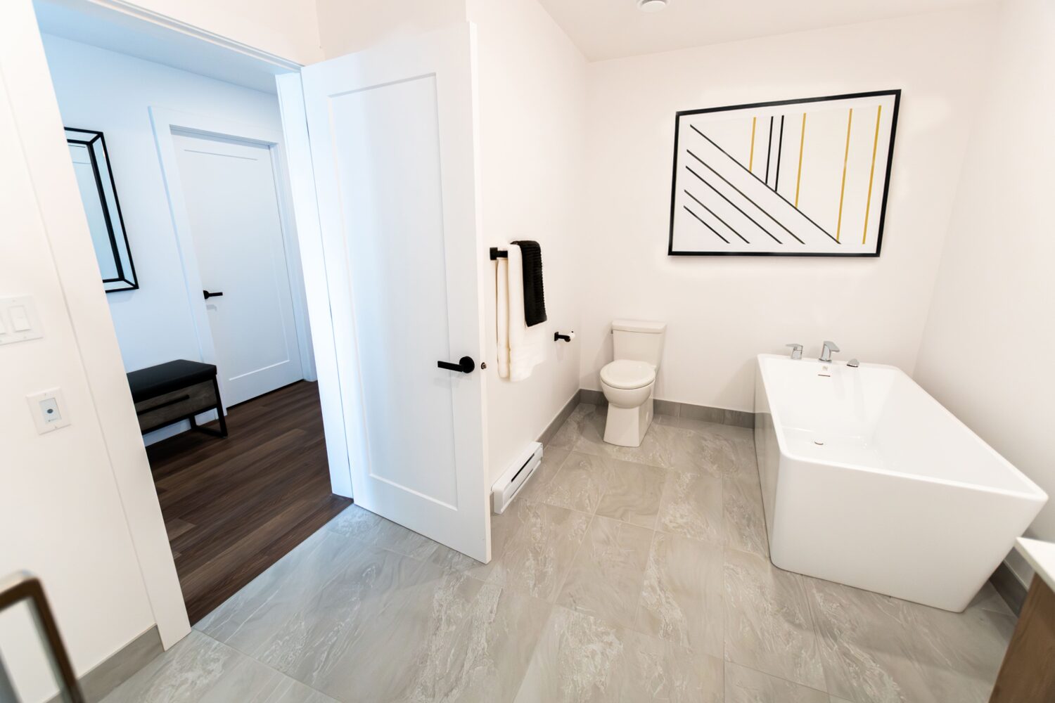 Vue d'une salle de bain moderne avec un bain autoportant blanc sur un plancher de tuiles grises. La pièce a des murs blancs, une toilette et un porte-serviettes. Un tableau abstrait avec des lignes noires et dorées est accroché au mur au-dessus de la baignoire. Une porte ouverte donne sur l'entrée de l'appartement avec un banc et un miroir.