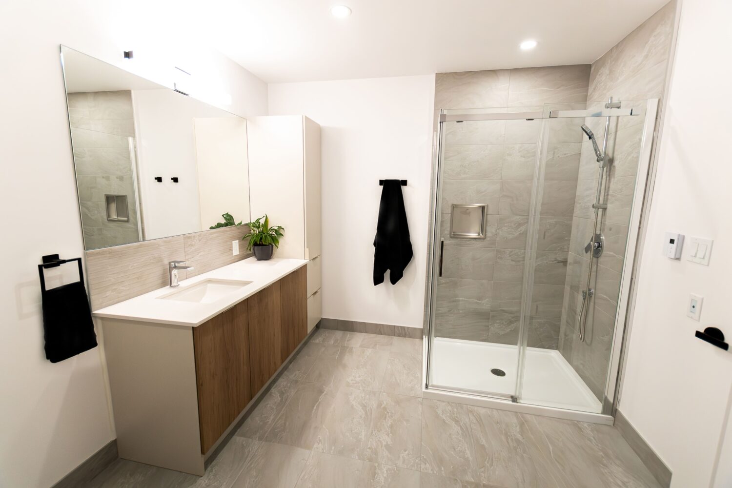 Vue d'une salle de bain moderne avec un grand meuble-lavabo en bois clair et blanc surmonté d'un comptoir blanc, d'un grand miroir et d'un lavabo encastré. La pièce a un plancher de tuiles grises et une grande douche en verre avec un pommeau de douche effet de pluie. Des porte-serviettes noirs et une petite plante en pot ajoutent des touches de décoration.