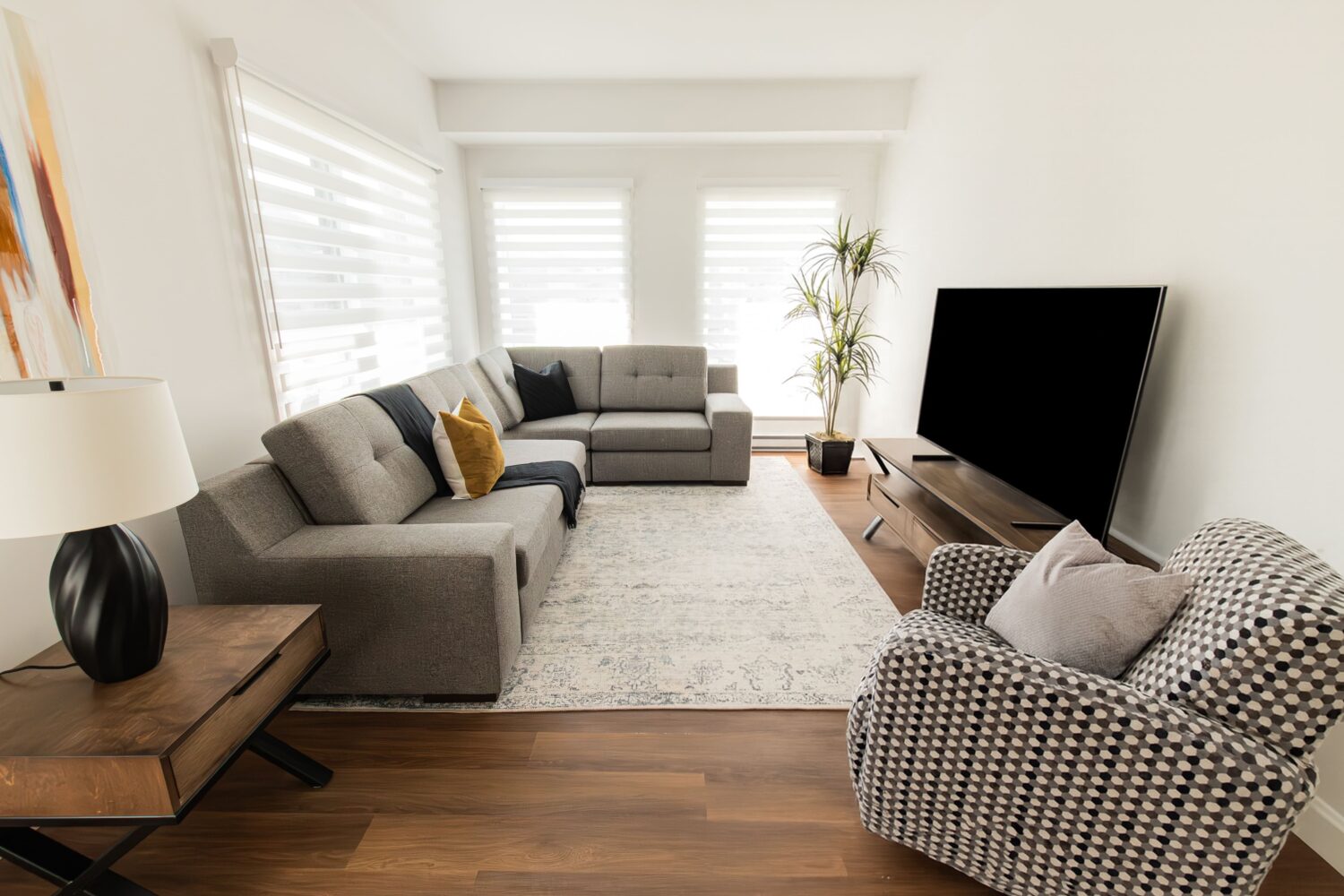 Vue d'un salon moderne et épuré avec un grand canapé d'angle en tissu gris et un fauteuil à motifs noir et blanc. La pièce dispose d'un plancher de bois, de murs blancs et d'un grand téléviseur à écran plat sur un meuble en bois foncé. Une lampe, une table d'appoint, un grand tapis, une plante en pot et des coussins d'appoint ajoutent une touche de confort.