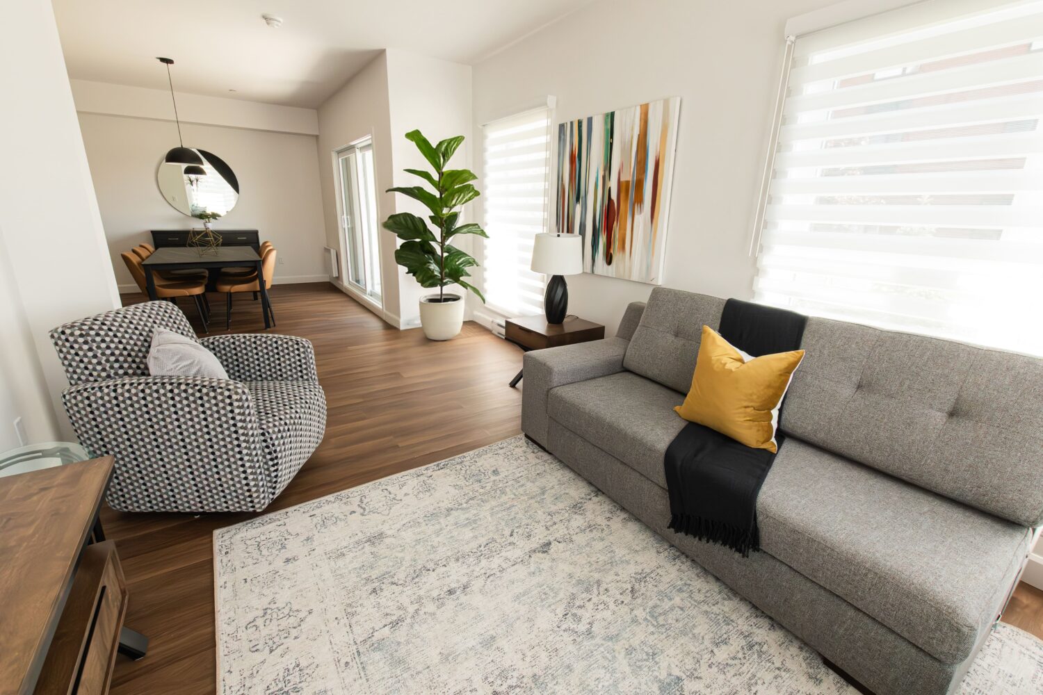Vue d'un salon à aire ouverte moderne qui s'étend vers une salle à manger. Le salon est meublé d'un canapé gris et d'un fauteuil à motifs noir et blanc, avec un grand tapis. Une œuvre d'art abstraite colorée est accrochée au-dessus du canapé. La salle à manger au fond est meublée d'une table, de chaises et d'un grand miroir rond sur le mur. Une grande plante en pot est placée près d'une porte patio.